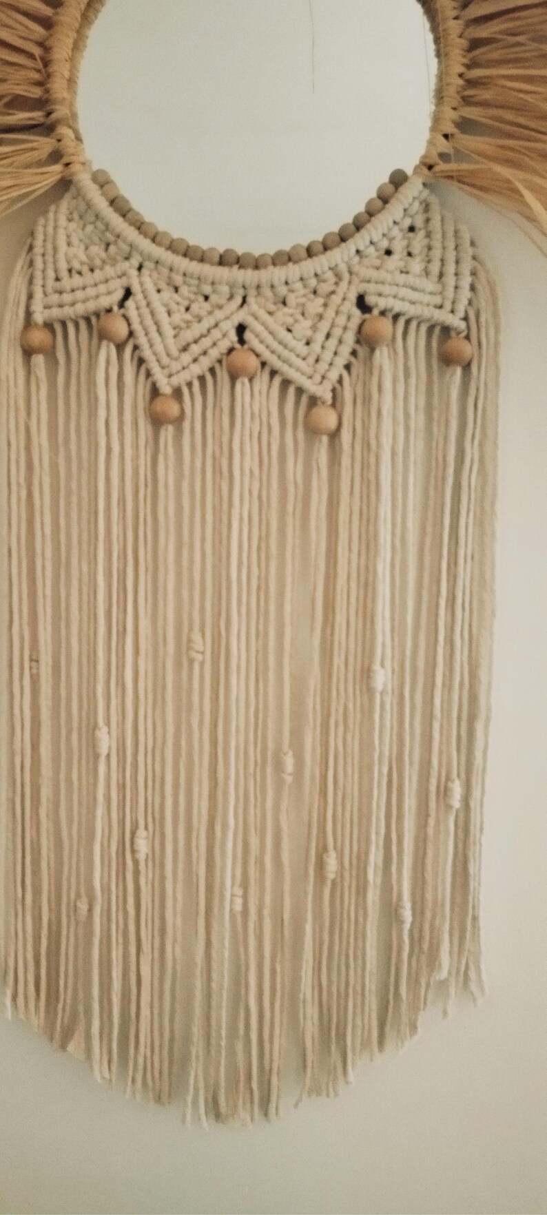 Puede incluir: Un tapiz de pared de macram&eacute; color crema con un dise&ntilde;o circular. La parte superior presenta un patr&oacute;n tejido con cuentas y detalles de madera. Largos hilos verticales de hilo cuelgan debajo, con cuentas de madera espaciadas a lo largo. Est&eacute;tica bohemia.