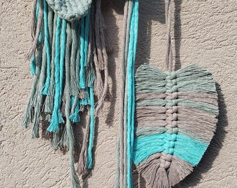 Op de afbeelding: Macramé wandhanger met een bloem- en bladerontwerp in grijstinten en teal. De bloem heeft een geometrisch midden en het blad is een gelaagd ontwerp. Bevestigd aan een houten deuvel.