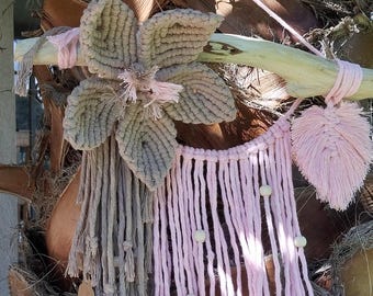 May include: A macrame wall hanging suspended from a light-colored wooden branch. It features a large, taupe-colored macrame flower with a pink center, a pink macrame leaf, and a section of vertical pink fringe adorned with small, round wooden beads.