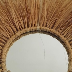 Miroir décoratif rond avec un cadre en raphia tissé naturel et un fond en macramé. Le miroir a un diamètre d'environ 30 cm et présente une palette de couleurs neutres façon bohème.