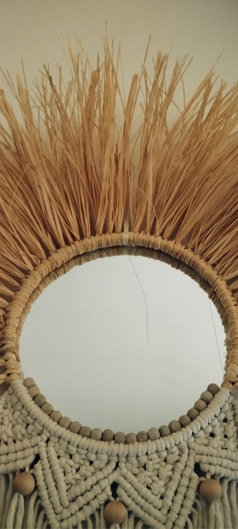Miroir décoratif rond avec un cadre en raphia tissé naturel et un fond en macramé. Le miroir a un diamètre d'environ 30 cm et présente une palette de couleurs neutres façon bohème.