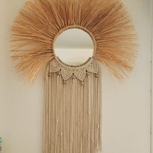 Puede incluir: Espejo de pared redondo con dise&ntilde;o de sol de fibra natural. El espejo est&aacute; rodeado por un marco de macram&eacute; beige, cuentas de madera y flecos colgantes. Una pieza decorativa para interiores bohemios o r&uacute;sticos.