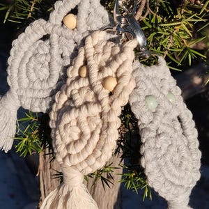 Puede incluir: Tres llaveros de búho de macramé en gris y crema, con ojos de cuentas de madera y borlas en la parte inferior. Los llaveros están sujetos a un cierre plateado.