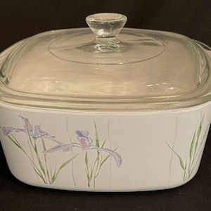 May include: A white square casserole dish with integrated side handles and a clear glass lid. The exterior of the dish features a delicate floral pattern of purple iris flowers and green stems painted against a clean white background surface.