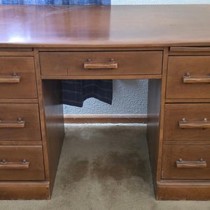 May include: A brown wooden desk featuring a rectangular top surface, a single central drawer, and two columns of three drawers each. Each of the seven drawers is equipped with a horizontal wooden handle centered on the front panel.