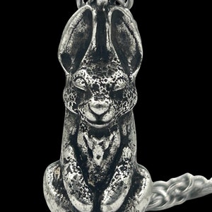 May include: A silver charm in the shape of a sitting rabbit with long ears. The charm is detailed with a textured surface and has a loop at the top for attaching to a chain or bracelet.