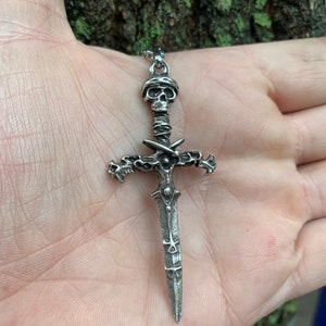 May include: A silver pendant necklace featuring a detailed skull and sword design. The sword has a cross guard with a skull at the top and a pointed blade.