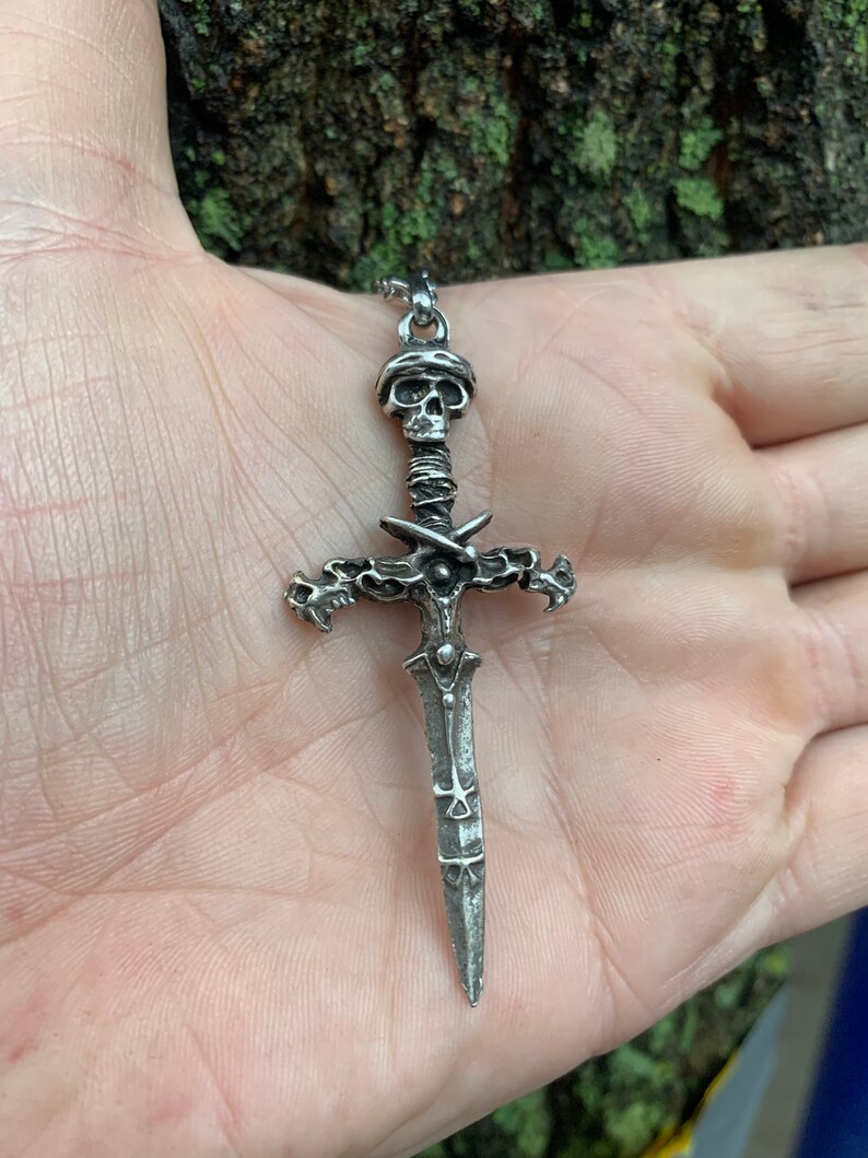 May include: A silver pendant necklace featuring a detailed skull and sword design. The sword has a cross guard with a skull at the top and a pointed blade.