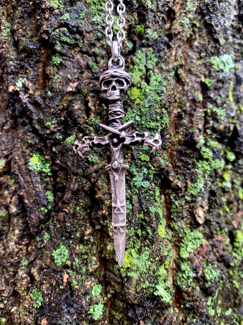 May include: A silver skull and sword pendant necklace. The pendant features a detailed skull with a wrapped sword handle and a cross-shaped hilt. The sword blade is decorated with intricate designs.