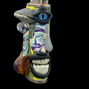 Może przedstawiać: Unikalna, artystyczna zapalniczka z motywem twarzy. Zapalniczka posiada metalową górę, niebieskie oko, uśmiechnięte usta oraz kolorowe, wirujące wzory w odcieniach niebieskiego, zielonego, żółtego i fioletowego. Projekt jest fantazyjny i przyciągający wzrok.