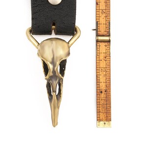 May include: A brass skull-shaped belt buckle with a black leather strap. The buckle is about 2 inches tall.