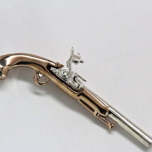 May include: A silver and gold-toned pendant necklace featuring a detailed replica of a pistol. The pendant is attached to a silver chain.