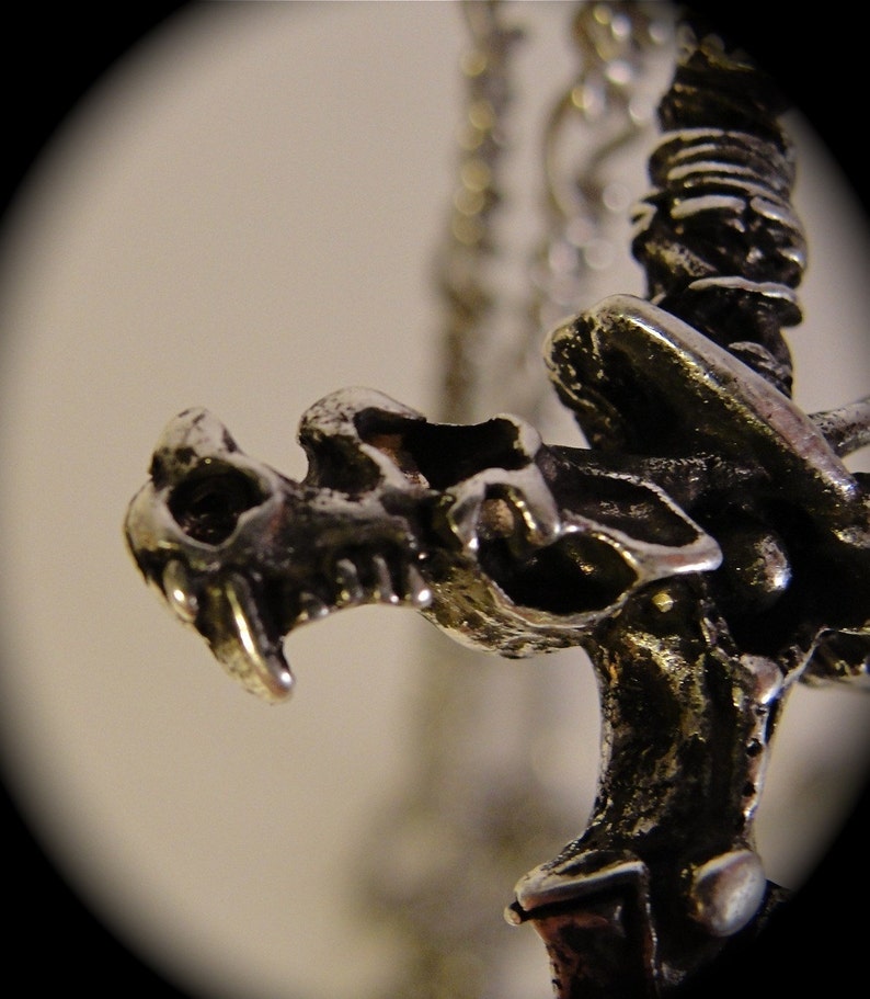May include: Close-up of a silver dragon pendant with intricate details. The dragon's head is facing forward with its mouth open, and its wings are spread out behind it.