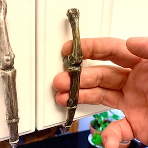 May include: A skeletal hand-shaped cabinet door pull in a brass finish. The pull is made to look like a bone with a textured surface.
