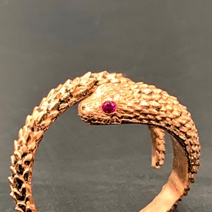 Puede incluir: Un anillo de serpiente dorado con un ojo de piedra preciosa rojo rubí. La serpiente está enroscada alrededor de la banda del anillo.