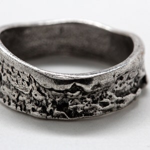 May include: A textured silver ring with a rugged, organic design. The ring's surface has a rough, uneven texture, creating a unique and weathered appearance. The ring is a simple band, perfect for everyday wear.