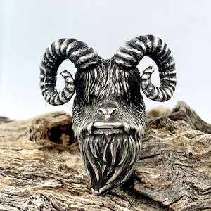 May include: A silver-colored pendant featuring a detailed sculpture of a goat's head with large horns and a beard. The pendant is on a wooden surface.