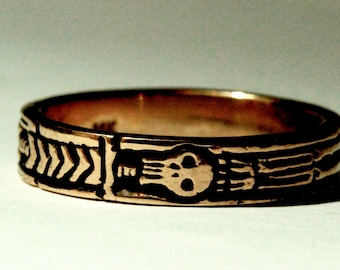 Könnte beinhalten: Ein goldener Ring mit einem Totenkopf- und Knochen-Design. Der Ring ist mit einem sich wiederholenden Muster von Pfeilen verziert.
