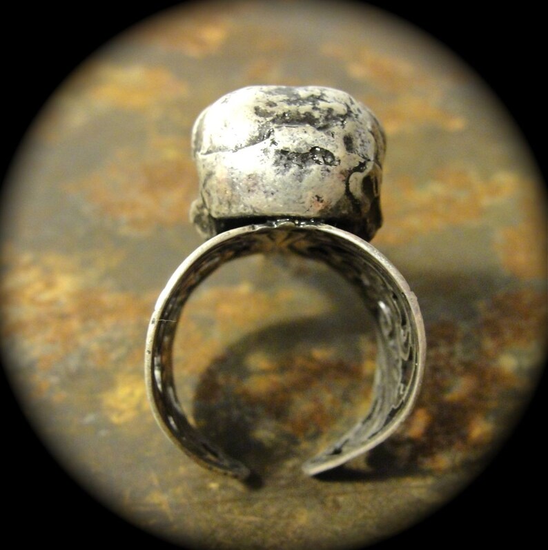 Puede incluir: Anillo plateado con un centro en forma de calavera. El anillo tiene un dise&ntilde;o abierto con intrincados patrones a lo largo de la banda. La calavera tiene una apariencia texturizada y desgastada.