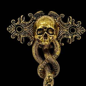 May include: A gold-toned metal hook featuring a skull and snake design. The skull has a detailed face and the snake is coiled around the skull.