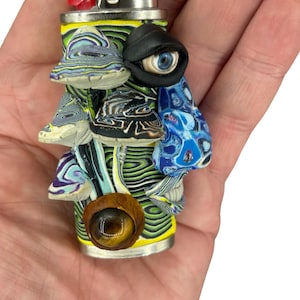 Może przedstawiać: Niestandardowo zdobiona zapalniczka BIC o unikalnym designie. Zapalniczka ma żółto-zielony wzór z wirami, ozdobiony rzeźbionymi kształtami przypominającymi grzyby w różnych kolorach i fakturach oraz dużym okiem. Dół ma drewniany akcent z brązowym kamieniem przypominającym oko.