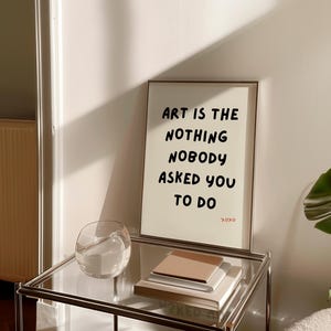 Op de afbeelding: Een ingelijst print met de tekst "Art is the nothing nobody asked you to do" op een witte achtergrond. De print staat op een glazen tafel met een glazen kom en een stapel boeken.