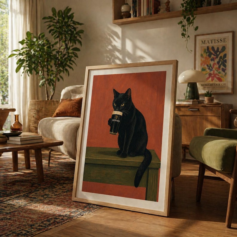 Puede incluir: Impresi&oacute;n art&iacute;stica enmarcada que presenta un gato negro sosteniendo una pinta de Guinness. El gato est&aacute; posado sobre una superficie verde contra un fondo rojo-naranja. La impresi&oacute;n est&aacute; en un marco de madera clara, agregando calidez.