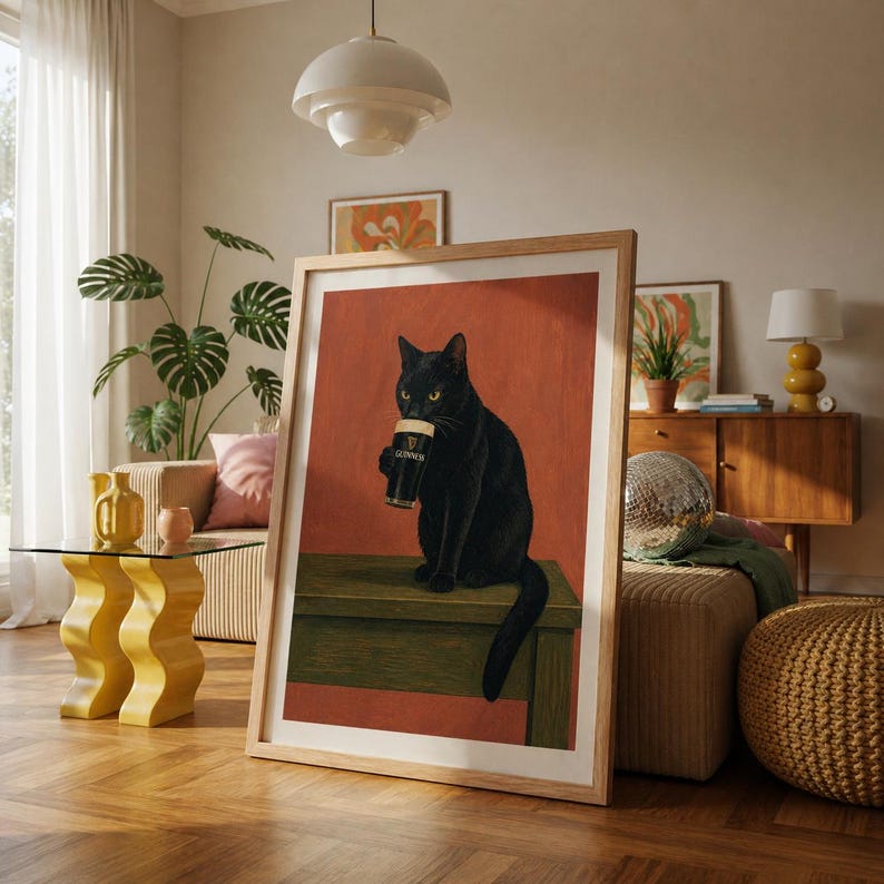 Puede incluir: Impresi&oacute;n art&iacute;stica enmarcada que muestra un gato negro bebiendo una pinta de Guinness. El gato est&aacute; sentado sobre una superficie verde, con un fondo rojo-anaranjado. La obra de arte se exhibe en una habitaci&oacute;n de dise&ntilde;o moderno.
