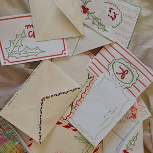 May include: A collection of Christmas-themed envelopes with various designs. Some envelopes feature holly and berry illustrations, while others have striped patterns and decorative borders. The color palette includes red, green, and white.