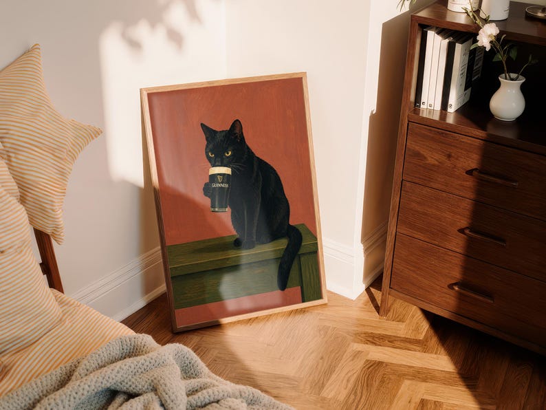 Puede incluir: Una impresi&oacute;n art&iacute;stica enmarcada que presenta un gato negro sosteniendo una pinta de Guinness. El gato, con ojos amarillos, est&aacute; sentado sobre una superficie verde contra un fondo rojo. La impresi&oacute;n est&aacute; en un marco de madera, agregando calidez a la obra.