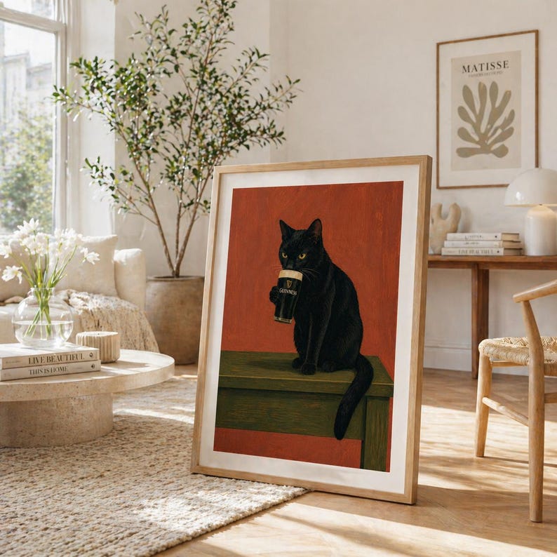 Puede incluir: Impresi&oacute;n enmarcada de un gato negro sosteniendo una pinta de Guinness. El gato est&aacute; sentado sobre una superficie verde contra un fondo rojo. La impresi&oacute;n est&aacute; en un marco de madera en una habitaci&oacute;n luminosa.
