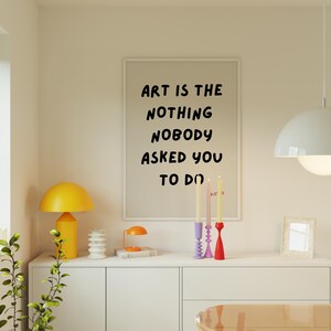Op de afbeelding: Een wit ingelijst print met de tekst "Art is the nothing nobody asked you to do" hangt aan een muur boven een witte commode met drie kandelaars en een gele lamp.