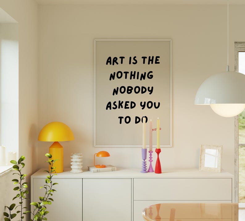 Op de afbeelding: Een wit ingelijst print met de tekst "Art is the nothing nobody asked you to do" hangt aan een muur boven een witte commode met drie kandelaars en een gele lamp.