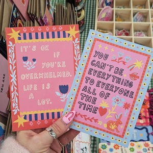 May include: Two colorful postcards with inspirational quotes. One pink card reads "It's ok if you're overwhelmed. Life is a lot." The other blue card says "You can't be everything to everyone all of the time."