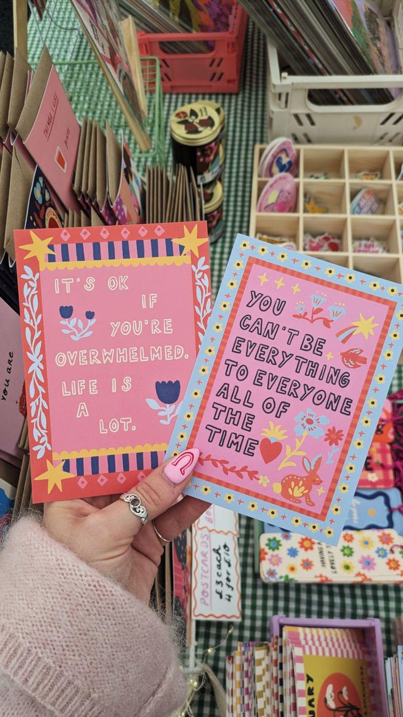 May include: Two colorful postcards with inspirational quotes. One pink card reads "It's ok if you're overwhelmed. Life is a lot." The other blue card says "You can't be everything to everyone all of the time."