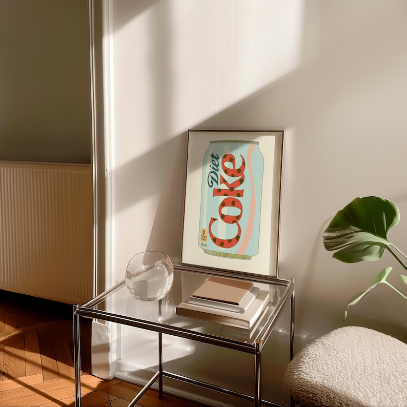 May include: A framed art print featuring a Diet Coke can design. The can is light blue with a red and pink swirl and the word "Diet Coke" in white. The print is displayed on a glass table with a clear glass and books.