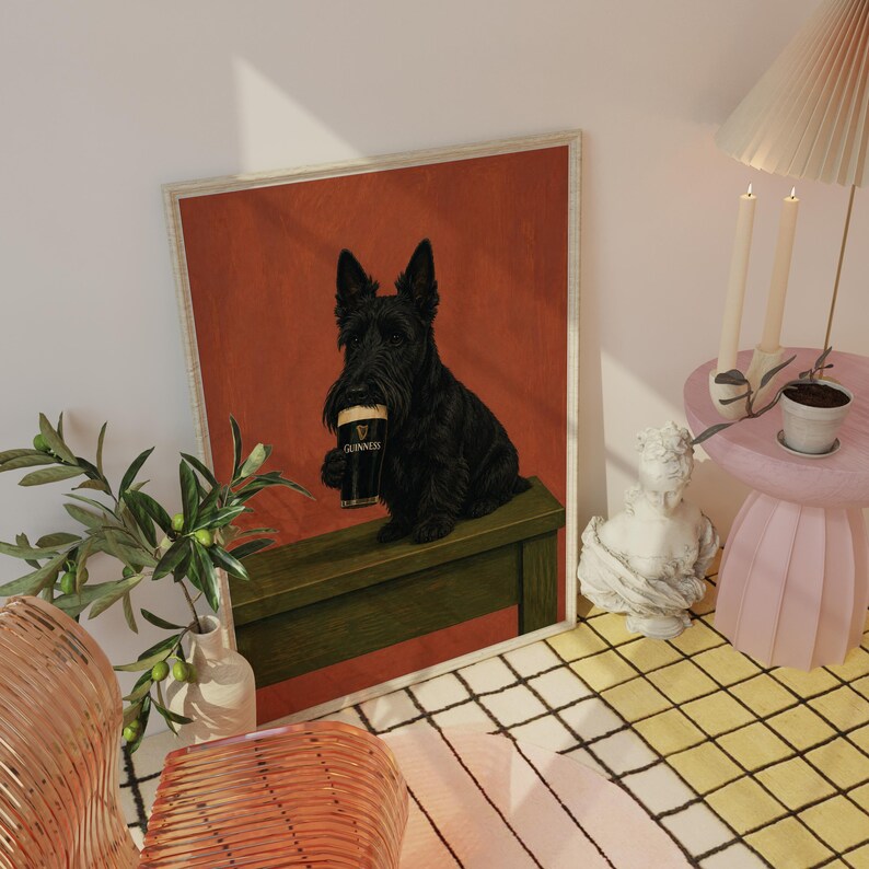 May include: A framed print featuring a black Scottish Terrier holding a Guinness pint. The dog is sitting on a green surface against a red background. The print is in a room with a lamp, candles, and a small table.