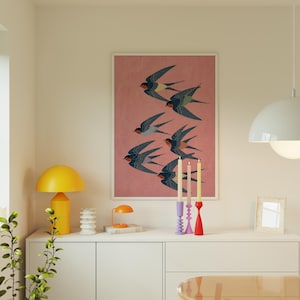 Puede incluir: Una impresi&oacute;n art&iacute;stica enmarcada que muestra una bandada de golondrinas en vuelo sobre un fondo rosa. Las aves est&aacute;n representadas en tonos azules, negros y rojos. La obra de arte se exhibe sobre un mueble blanco con objetos decorativos.