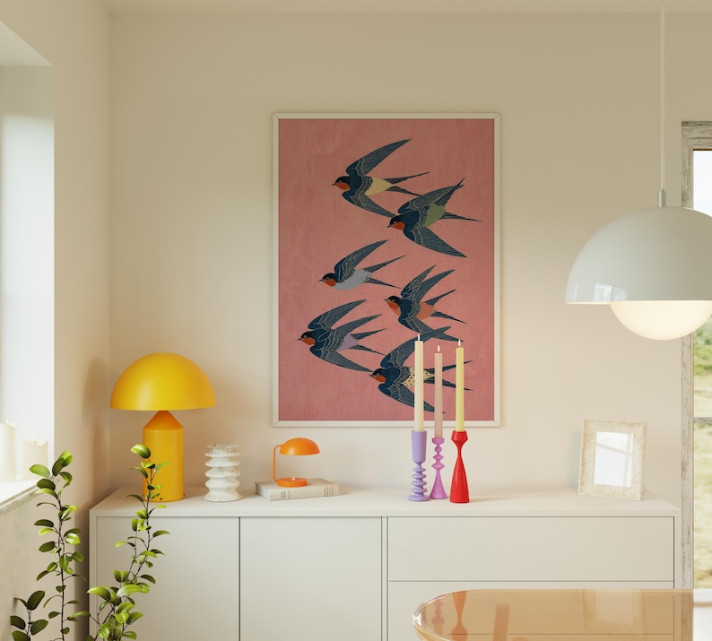 Puede incluir: Una impresi&oacute;n art&iacute;stica enmarcada que muestra una bandada de golondrinas en vuelo sobre un fondo rosa. Las aves est&aacute;n representadas en tonos azules, negros y rojos. La obra de arte se exhibe sobre un mueble blanco con objetos decorativos.