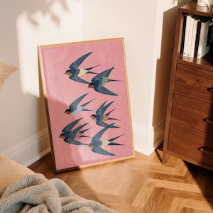 Puede incluir: Una impresi&oacute;n art&iacute;stica enmarcada que presenta una bandada de golondrinas estilizadas en vuelo. Las aves est&aacute;n representadas en tonos de azul, verde azulado y rojo sobre un fondo rosa. La obra de arte se muestra en un marco de madera, apoyada contra una pared.