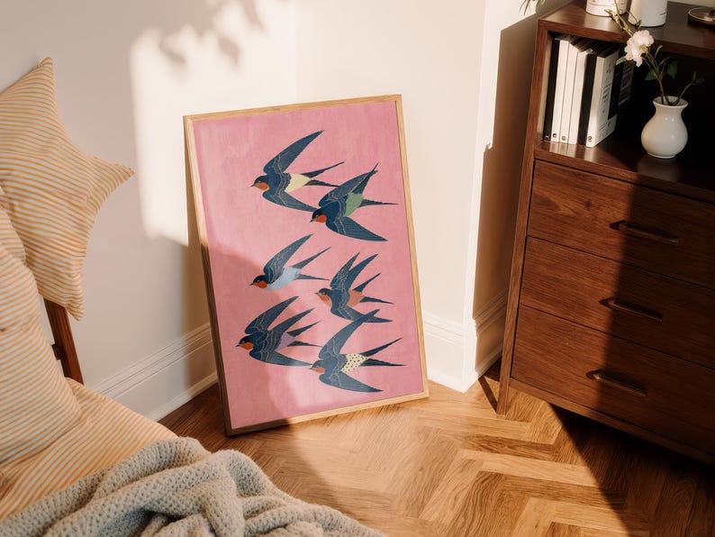Puede incluir: Una impresi&oacute;n art&iacute;stica enmarcada que presenta una bandada de golondrinas estilizadas en vuelo. Las aves est&aacute;n representadas en tonos de azul, verde azulado y rojo sobre un fondo rosa. La obra de arte se muestra en un marco de madera, apoyada contra una pared.