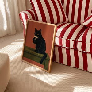 Puede incluir: Una impresi&oacute;n art&iacute;stica enmarcada que presenta un gato negro sosteniendo una pinta de Guinness. El gato est&aacute; posado sobre una superficie verde contra un fondo rojo anaranjado. El marco es de color madera clara. La impresi&oacute;n se exhibe cerca de un sof&aacute; a rayas rojas y blancas.