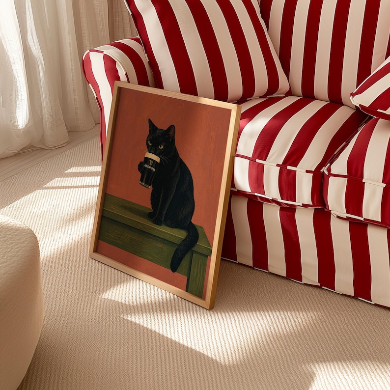 Puede incluir: Una impresi&oacute;n art&iacute;stica enmarcada que presenta un gato negro sosteniendo una pinta de Guinness. El gato est&aacute; posado sobre una superficie verde contra un fondo rojo anaranjado. El marco es de color madera clara. La impresi&oacute;n se exhibe cerca de un sof&aacute; a rayas rojas y blancas.