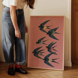 Puede incluir: Un cuadro enmarcado con una bandada de golondrinas en vuelo. Las aves tienen alas azules, negras y blancas con detalles rojos, sobre un fondo rosa. La obra de arte est&aacute; en un marco de madera.