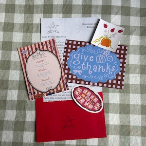 May include: A collection of stationery items, including a red envelope, a menu card, and cards with illustrations and text. The cards feature a star logo, a pumpkin, and the phrase "give thanks."