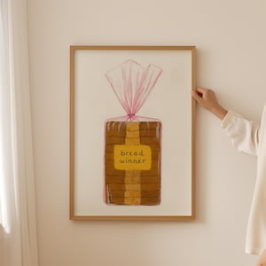 Op de afbeelding: Een ingelijste kunstdruk met een brood in een roze plastic zak. Het brood heeft een geel etiket met de tekst "bread winner". Het kunstwerk zit in een houten lijst.