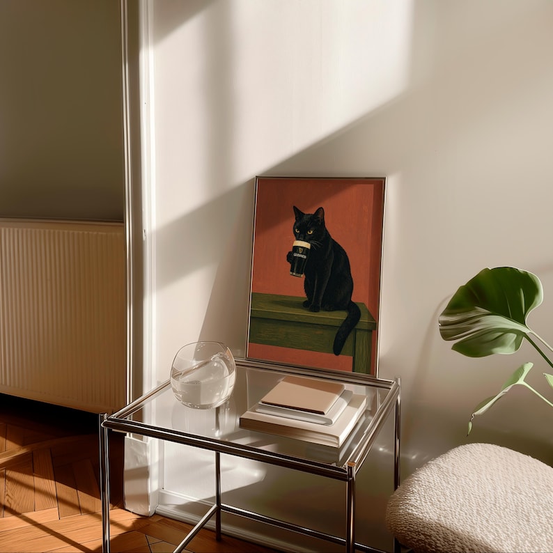 Puede incluir: Una impresi&oacute;n art&iacute;stica enmarcada que presenta un gato negro sosteniendo una pinta de cerveza negra. El gato est&aacute; sentado en un banco verde sobre un fondo rojo. La impresi&oacute;n se exhibe en una mesa auxiliar de vidrio y metal, con un cuenco de vidrio transparente y libros apilados.