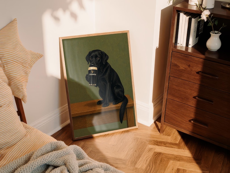 Pu&ograve; includere: Stampa incorniciata con un Labrador Retriever nero che tiene in mano una pinta di Guinness. Il cane &egrave; seduto su una panca di legno su uno sfondo verde. La stampa &egrave; esposta in una stanza con mobili in legno e pavimento a motivi.