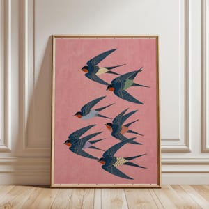 Puede incluir: Una impresi&oacute;n art&iacute;stica enmarcada que representa una bandada de golondrinas en vuelo. Las aves tienen alas azul marino, gargantas rojas y detalles en amarillo, verde y blanco. El fondo es rosa suave y el marco es marr&oacute;n claro.