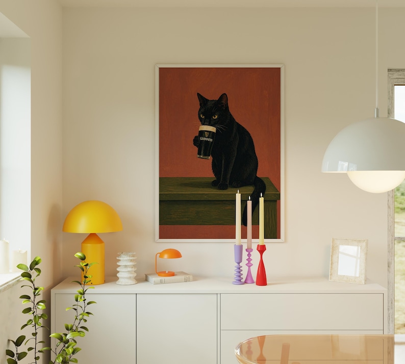 Puede incluir: Una impresi&oacute;n art&iacute;stica enmarcada representa un gato negro sosteniendo una pinta de Guinness. El gato est&aacute; sentado sobre una superficie verde contra un fondo rojo-naranja. La imagen est&aacute; enmarcada en blanco. La escena se complementa con candelabros coloridos.